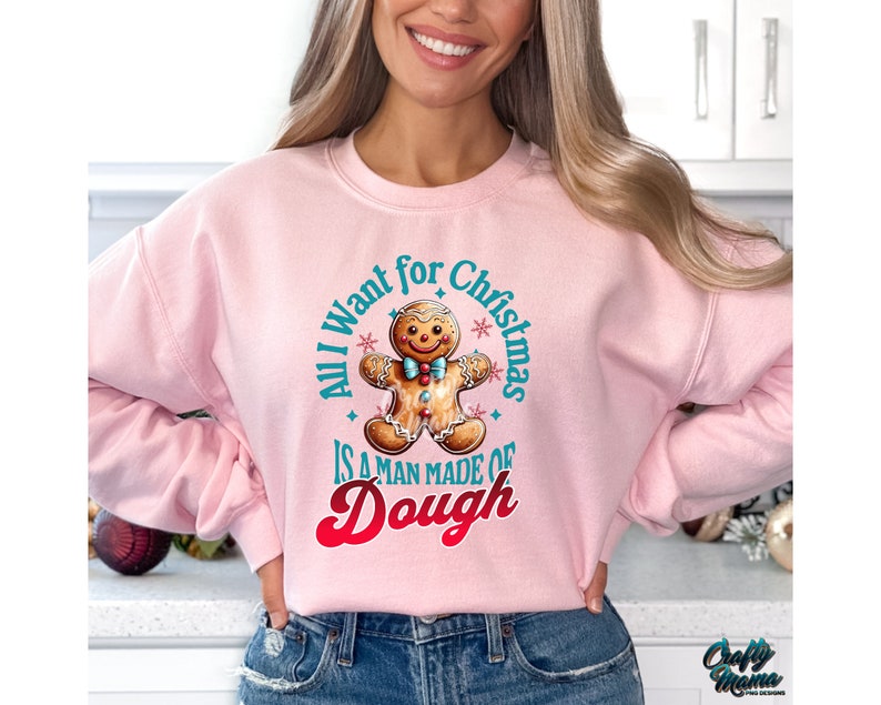 May include: A light pink sweatshirt with the text "All I want for Christmas is a man made of dough" and a gingerbread man graphic. The graphic is in shades of brown, blue, and red. The sweatshirt is a crew neck style.
