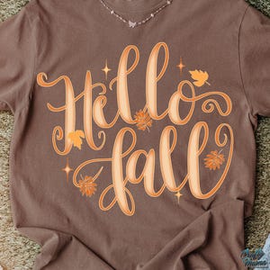 Puede incluir: Una camiseta marr&oacute;n con las palabras "Hello Fall" en letras grandes y estilizadas de color naranja. El dise&ntilde;o incluye hojas naranjas y acentos de estrellas, evocando un tema oto&ntilde;al. Se ve un delicado collar en el escote.