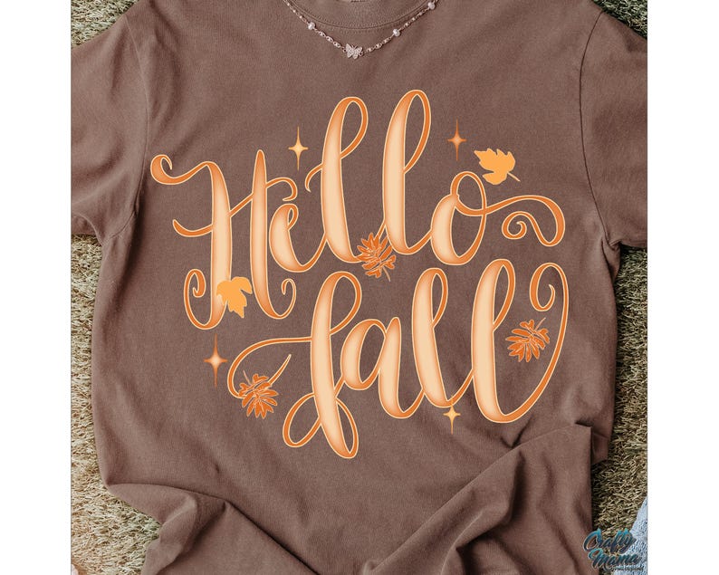 Puede incluir: Una camiseta marr&oacute;n con las palabras "Hello Fall" en letras grandes y estilizadas de color naranja. El dise&ntilde;o incluye hojas naranjas y acentos de estrellas, evocando un tema oto&ntilde;al. Se ve un delicado collar en el escote.