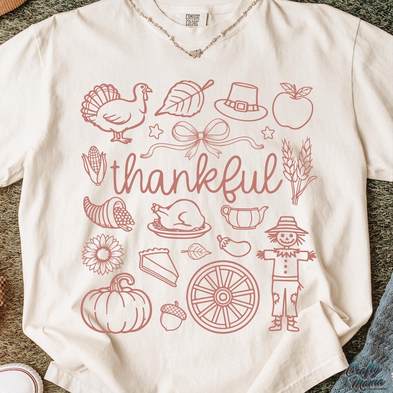 May include: Cream-colored t-shirt with a pink graphic design featuring Thanksgiving-themed illustrations. The word "thankful" is written in cursive. The design includes a turkey, leaf, hat, apple, bow, cornucopia, pie, pumpkin, and scarecrow.
