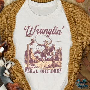 May include: White t-shirt with a graphic featuring a cowboy riding a horse, roping a raccoon, and the text "Wranglin' Feral Children." The design is in shades of brown and purple, with desert scenery. The shirt is displayed on a mustard-colored cardigan.