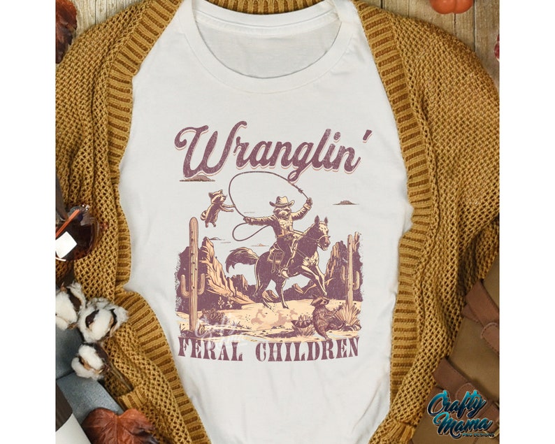 May include: White t-shirt with a graphic featuring a cowboy riding a horse, roping a raccoon, and the text "Wranglin' Feral Children." The design is in shades of brown and purple, with desert scenery. The shirt is displayed on a mustard-colored cardigan.