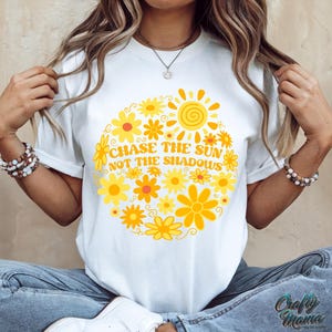 May include: White t-shirt with a circular floral design in yellow and orange hues, featuring a sun graphic and the text "CHASE THE SUN NOT THE SHADOWS". The shirt is a casual wear item.