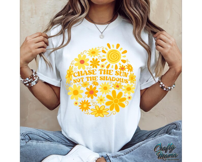 May include: White t-shirt with a circular floral design in yellow and orange hues, featuring a sun graphic and the text "CHASE THE SUN NOT THE SHADOWS". The shirt is a casual wear item.