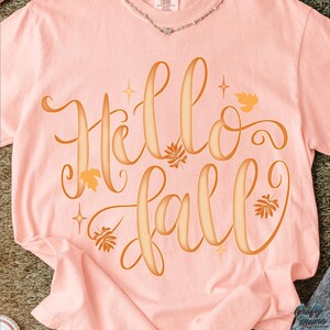 Puede incluir: Una camiseta rosa claro con las palabras "Hello Fall" en naranja c&aacute;lido. El dise&ntilde;o incluye hojas y estrellas, perfecto para el oto&ntilde;o. La camiseta est&aacute; hecha de un material suave.
