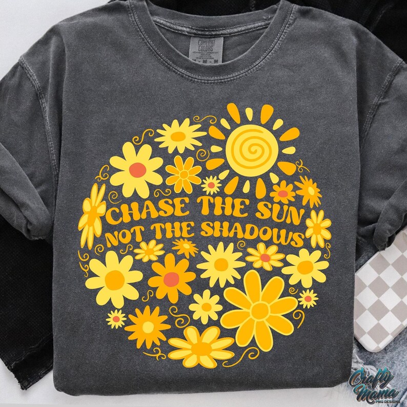 May include: Dark gray sweatshirt with a floral design in yellow and orange. The design includes a sun and the text "CHASE THE SUN NOT THE SHADOWS". The sweatshirt has long sleeves and a crew neck.