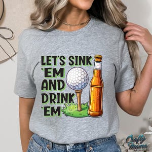 Pode incluir: Camiseta cinza com o texto "Let's Sink 'Em and Drink 'Em" em verde e preto. O design inclui uma bola de golfe em um tee e uma garrafa de cerveja. A camiseta &eacute; de estilo casual.