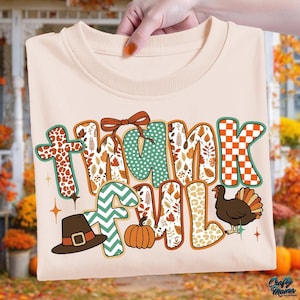 May include: A beige t-shirt with the words "Thankful" in a colorful, fall-themed design. The letters feature patterns like leopard print, polka dots, and autumn leaves. A turkey, pumpkin, and pilgrim hat are also included.