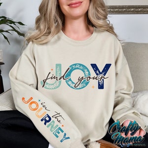 May include: A light beige sweatshirt with the text "find your JOY" in blue, orange, and teal lettering with a distressed effect. The left sleeve has the text "in the JOURNEY" in a rainbow color scheme.