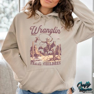 May include: Beige hoodie with a front pocket and drawstring hood. The graphic features a cowboy on a horse roping a raccoon, with the words "Wranglin' Feral Children" in a purple font. The Crafty Mama logo is in the bottom right corner.