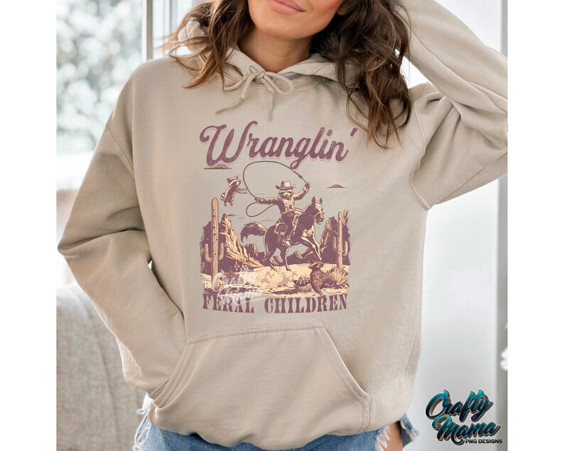 May include: Beige hoodie with a front pocket and drawstring hood. The graphic features a cowboy on a horse roping a raccoon, with the words "Wranglin' Feral Children" in a purple font. The Crafty Mama logo is in the bottom right corner.