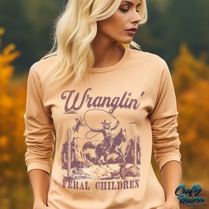 May include: A long-sleeved, tan-colored shirt featuring a vintage-style graphic of a cowboy roping a steer, with the words "Wranglin' Feral Children" in a purple, distressed font. The shirt has a crew neck and a relaxed fit.