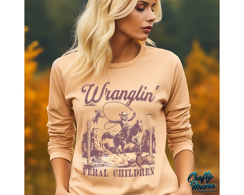 May include: A long-sleeved, tan-colored shirt featuring a vintage-style graphic of a cowboy roping a steer, with the words "Wranglin' Feral Children" in a purple, distressed font. The shirt has a crew neck and a relaxed fit.