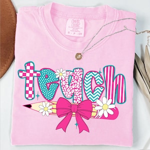 May include: Pink t-shirt with the word "teach" in colorful patterned letters. A yellow pencil with a pink bow and white daisies is below the word.
