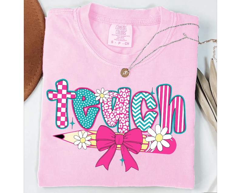 May include: Pink t-shirt with the word "teach" in colorful patterned letters. A yellow pencil with a pink bow and white daisies is below the word.