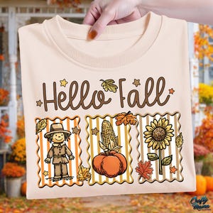 Puede incluir: Una camiseta beige con las palabras "Hello Fall" en escritura marrón. Debajo del texto hay tres imágenes rectangulares: un espantapájaros, una calabaza con maíz y un girasol. La camiseta está sujeta por una mano.