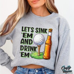 Pode incluir: Camisola cinzenta com o texto "LET'S SINK 'EM AND DRINK 'EM" em preto e verde. O gr&aacute;fico inclui uma bola de golfe num tee, uma garrafa de cerveja e relva verde. A camisola &eacute; de estilo casual.