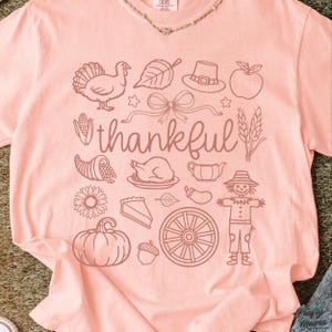 May include: A light pink t-shirt with a Thanksgiving-themed graphic. The design includes a turkey, leaf, hat, apple, bow, cornucopia, roasted turkey, sunflower, pumpkin, pie, and the word "thankful".
