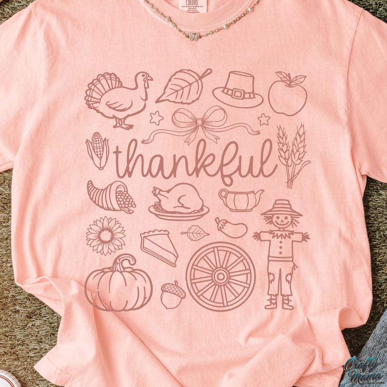 May include: A light pink t-shirt with a Thanksgiving-themed graphic. The design includes a turkey, leaf, hat, apple, bow, cornucopia, roasted turkey, sunflower, pumpkin, pie, and the word "thankful".