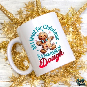 May include: White ceramic mug with a handle, featuring a gingerbread man illustration and the text "All I want for Christmas is a man made of dough" in teal and red. The mug is surrounded by gold star-shaped decorations.