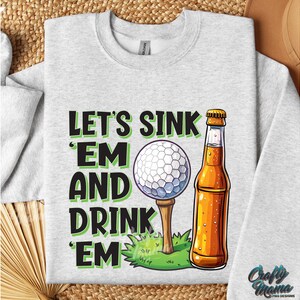 Pode incluir: Camisola cinza claro com um gr&aacute;fico de uma bola de golfe num tee, uma garrafa de cerveja e o texto "LET'S SINK 'EM AND DRINK 'EM". O design &eacute; em preto e verde. A camisola tem gola redonda e mangas compridas.