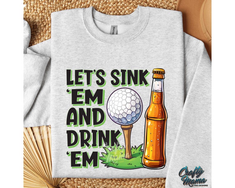 Pode incluir: Camisola cinza claro com um gr&aacute;fico de uma bola de golfe num tee, uma garrafa de cerveja e o texto "LET'S SINK 'EM AND DRINK 'EM". O design &eacute; em preto e verde. A camisola tem gola redonda e mangas compridas.