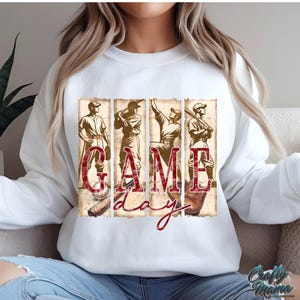 K&ouml;nnte beinhalten: Wei&szlig;es Sweatshirt mit einem Baseball-Motiv. Die Grafik zeigt vier Baseballspieler und die Worte "GAME DAY" in einem Vintage-Stil. Das Design verwendet Braun- und Rott&ouml;ne und erzeugt einen klassischen Sport-Look.