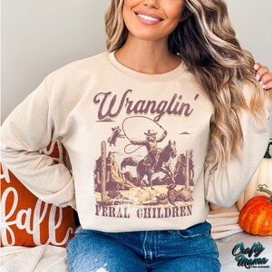 May include: Beige sweatshirt with a western-themed graphic. The graphic features a cowboy on a horse roping, with the words "Wranglin' Feral Children" in a vintage font. The sweatshirt has long sleeves and a crew neck.