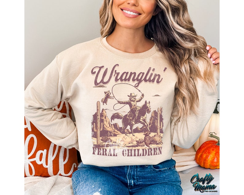 May include: Beige sweatshirt with a western-themed graphic. The graphic features a cowboy on a horse roping, with the words "Wranglin' Feral Children" in a vintage font. The sweatshirt has long sleeves and a crew neck.