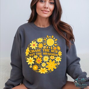 May include: Dark gray sweatshirt with a floral sun design. The design features yellow and orange flowers surrounding a sun graphic, with the text "CHASE THE SUN NOT THE SHADOWS". The sweatshirt is a crew neck style.