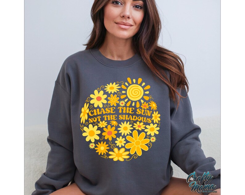 May include: Dark gray sweatshirt with a floral sun design. The design features yellow and orange flowers surrounding a sun graphic, with the text "CHASE THE SUN NOT THE SHADOWS". The sweatshirt is a crew neck style.