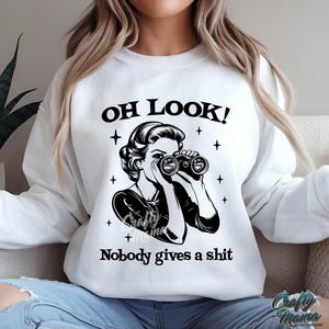 K&ouml;nnte beinhalten: Wei&szlig;es Sweatshirt mit dem Text "OH LOOK!" &uuml;ber einer Schwarz-Wei&szlig;-Illustration einer Frau mit Fernglas. Unter der Illustration steht der Text "Nobody gives a shit". Das Sweatshirt hat einen Rundhalsausschnitt.