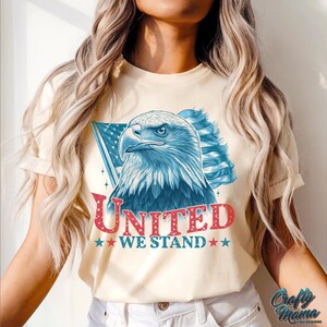 May include: A cream-coloured t-shirt featuring a graphic of a bald eagle with red, white and blue American flag wings. The text "UNITED WE STAND" is printed in red and blue below the eagle.