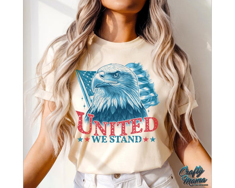 May include: A cream-coloured t-shirt featuring a graphic of a bald eagle with red, white and blue American flag wings. The text "UNITED WE STAND" is printed in red and blue below the eagle.