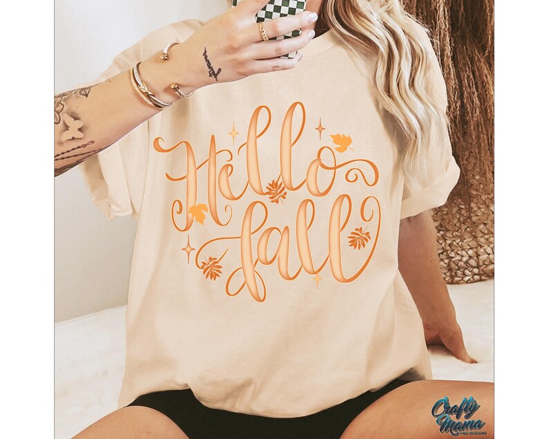 Puede incluir: Camiseta color crema con la frase "Hello Fall" en escritura naranja. El dise&ntilde;o incluye hojas y estrellas naranjas. Es una prenda de vestir casual y de temporada.
