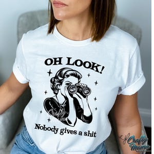 K&ouml;nnte beinhalten: Wei&szlig;es T-Shirt mit dem Text "OH LOOK!" &uuml;ber einer Schwarz-Wei&szlig;-Illustration einer Frau, die durch ein Fernglas blickt. Unter der Illustration steht der Text "Nobody gives a shit".