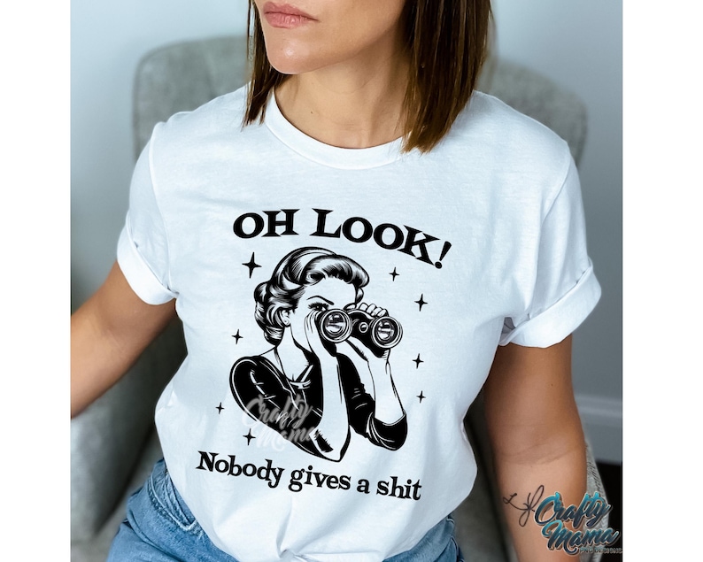 K&ouml;nnte beinhalten: Wei&szlig;es T-Shirt mit dem Text "OH LOOK!" &uuml;ber einer Schwarz-Wei&szlig;-Illustration einer Frau, die durch ein Fernglas blickt. Unter der Illustration steht der Text "Nobody gives a shit".