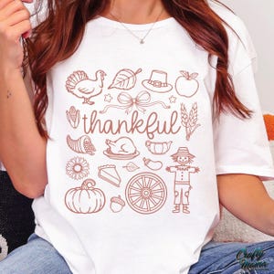 May include: White t-shirt with a brown graphic design featuring Thanksgiving-themed illustrations, including a turkey, leaf, hat, apple, and the word "thankful". The design also includes a bow, stars, cornucopia, and a scarecrow.