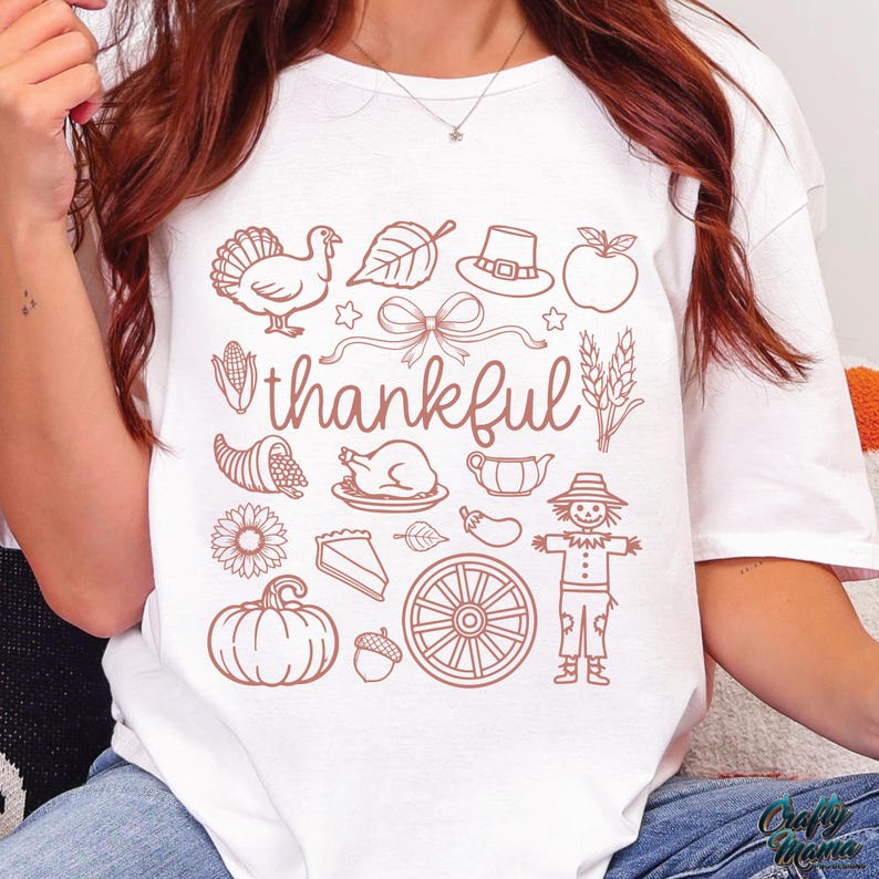 May include: White t-shirt with a brown graphic design featuring Thanksgiving-themed illustrations, including a turkey, leaf, hat, apple, and the word "thankful". The design also includes a bow, stars, cornucopia, and a scarecrow.