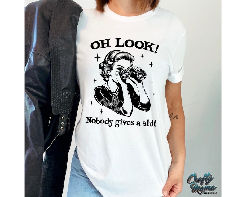 K&ouml;nnte beinhalten: Wei&szlig;es T-Shirt mit einem schwarzen Grafik einer Frau, die durch ein Fernglas blickt, mit dem Text "OH LOOK! Nobody gives a shit." Eine schwarze Lederjacke ist zu sehen.