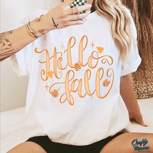 Puede incluir: Camiseta blanca con la frase "Hello Fall" en escritura naranja, adornada con hojas y estrellas. La camiseta es de estilo casual, de manga corta, perfecta para el oto&ntilde;o. El logo "Crafty Mama" es visible en la parte inferior derecha.