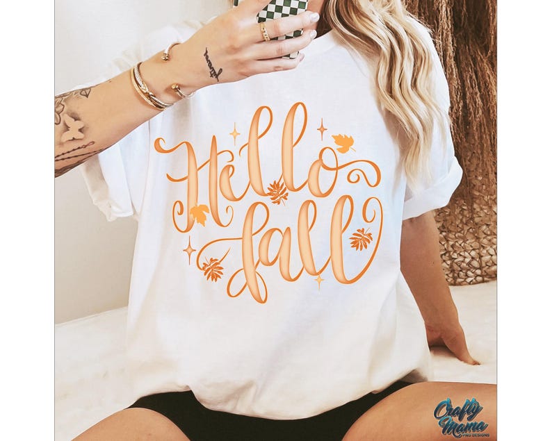 Puede incluir: Camiseta blanca con la frase "Hello Fall" en escritura naranja, adornada con hojas y estrellas. La camiseta es de estilo casual, de manga corta, perfecta para el oto&ntilde;o. El logo "Crafty Mama" es visible en la parte inferior derecha.
