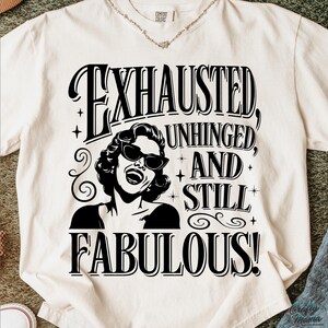 May include: Cream-coloured t-shirt with a black and white graphic of a woman in sunglasses, and the text "EXHAUSTED, UNHINGED, AND STILL FABULOUS!". The shirt is made of a soft material.