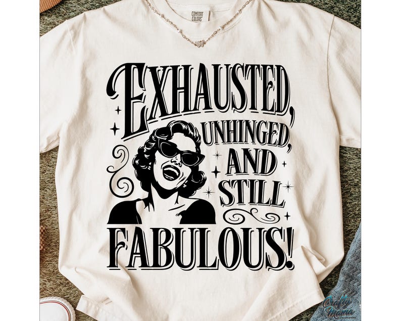 May include: Cream-coloured t-shirt with a black and white graphic of a woman in sunglasses, and the text "EXHAUSTED, UNHINGED, AND STILL FABULOUS!". The shirt is made of a soft material.