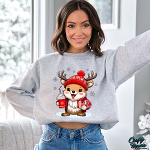 May include: A gray sweatshirt with a cute cartoon reindeer wearing a red hat and holding a red cup. The reindeer is surrounded by snowflakes.
