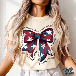 May include: A cream-colored t-shirt with a red, white, and blue bow design. The bow is surrounded by the text "Land of the Free * America * Land of the Free * America * Land of the Free * America * Land of the Free * America".