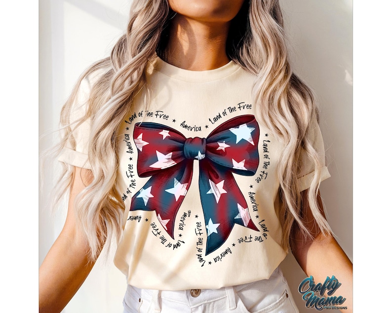 May include: A cream-colored t-shirt with a red, white, and blue bow design. The bow is surrounded by the text "Land of the Free * America * Land of the Free * America * Land of the Free * America * Land of the Free * America".