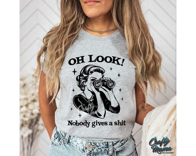 K&ouml;nnte beinhalten: Hellgraues T-Shirt mit dem Text "OH LOOK!" &uuml;ber einer schwarz-wei&szlig;en Grafik einer Frau mit Fernglas. Darunter steht der Text "Nobody gives a shit".