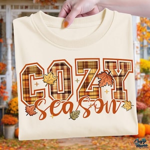 Może przedstawiać: Kremowa koszulka z długim rękawem z napisem "COZY season" w jesiennym motywie. Litery są obrysowane na brązowo z wzorem w kratę i ozdobione jesiennymi liśćmi i gwiazdkami.
