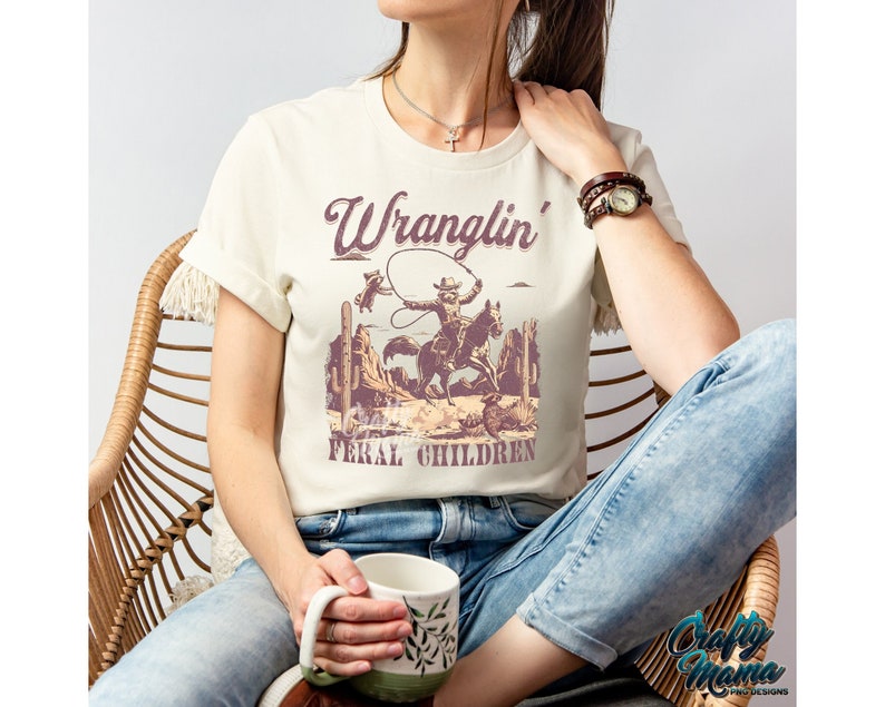 May include: Cream-colored t-shirt featuring a vintage-style graphic of a cowboy on a horse roping a steer, with the text "Wranglin' Feral Children" in a distressed font. The shirt is worn by a person sitting in a wicker chair.